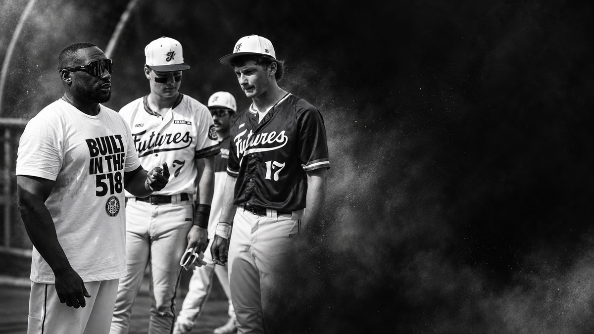 Black and white photo of four baseball players walking together on a field, with one player wearing a white 'Futures' jersey and another wearing a dark 'Futures' jersey with the number 17. A coach or staff member on the left wears a white t-shirt with the text 'BUILT IN THE 518'.