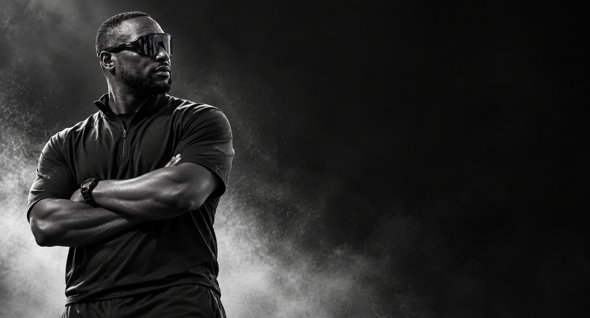 A black and white portrait of a man wearing dark sunglasses, a dark short-sleeve collared shirt, and a watch, standing with arms crossed against a smoky dark background.